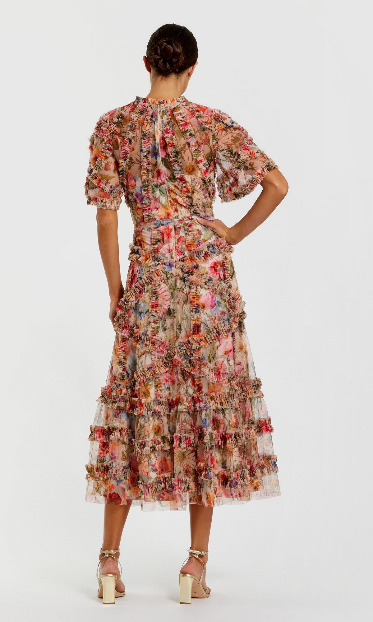 A woman faces away, wearing the Mac Duggal 8010 short-sleeve wedding-guest dress with ruffles and floral print, paired with white heeled sandals against a plain background.